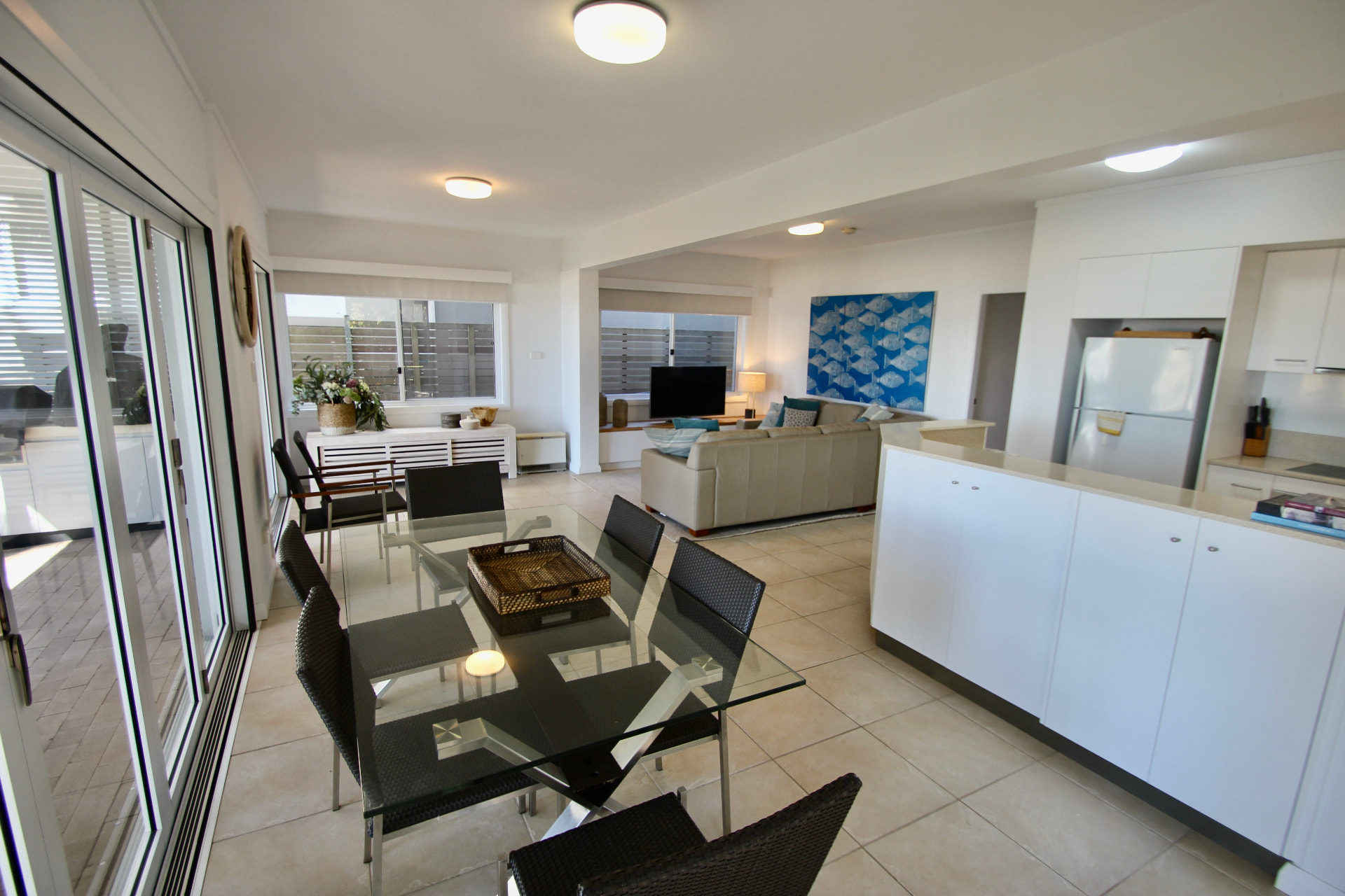Downstairs open-plan living and dining area with glass dining table, lounge and white kitchen, sliding doors to the terrace