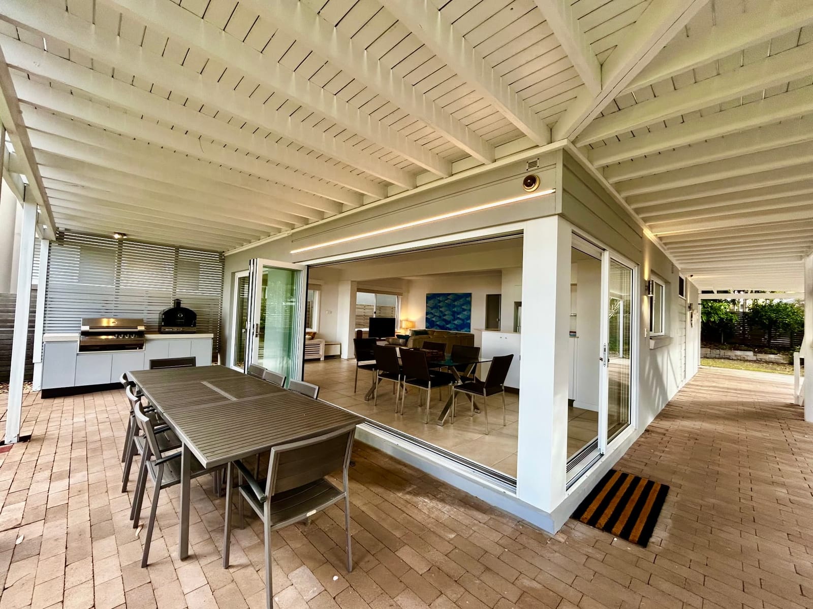 Downstairs covered terrace with long outdoor dining table, built-in BBQ and pizza oven, and bifold doors open to the living and dining room