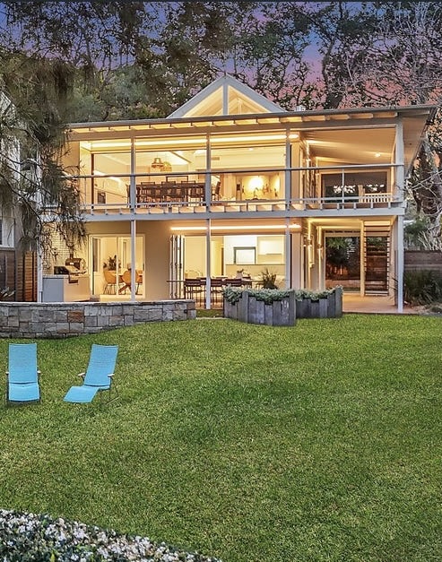 The Gem at dusk — two-storey beachfront house glowing warm against the trees, viewed from the garden