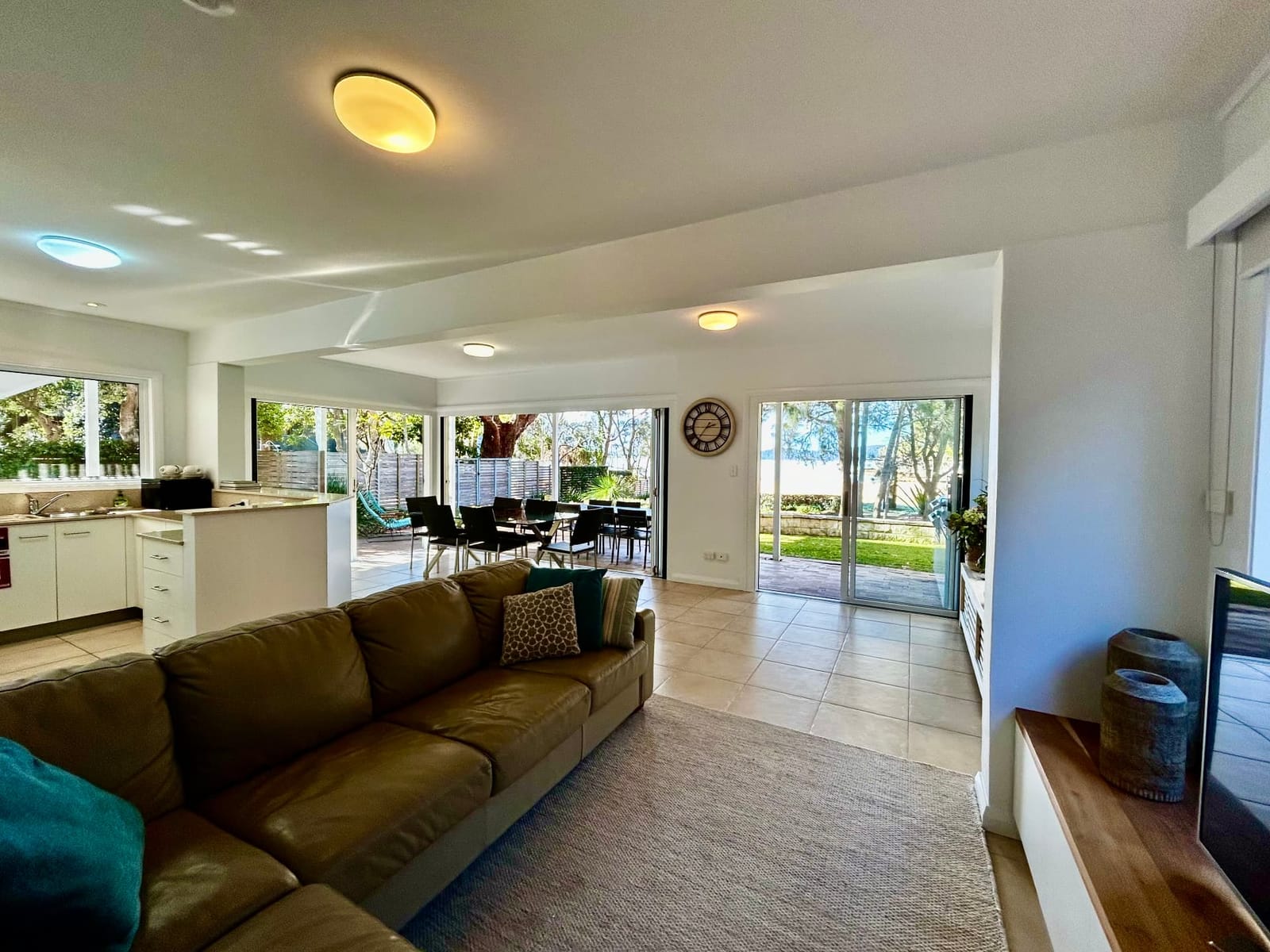 Downstairs lounge with leather sofa looking through the open-plan kitchen and dining area to the terrace, lawn and Pearl Beach beyond