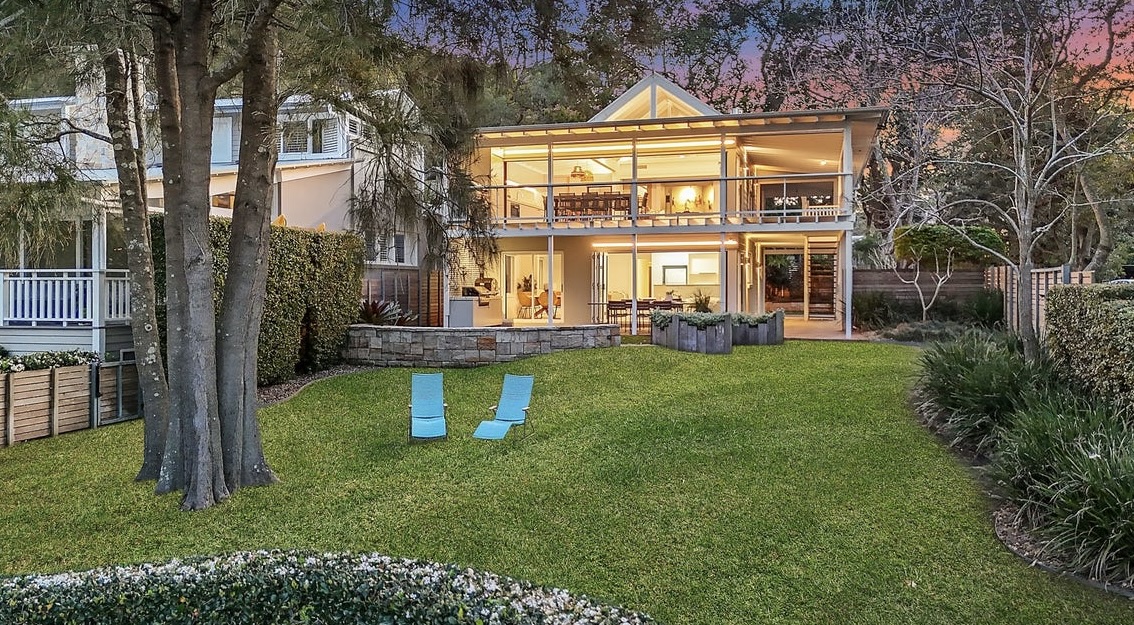 View of the house from the backyard at dusk — lawn, loungers and the lit interiors opening onto the garden