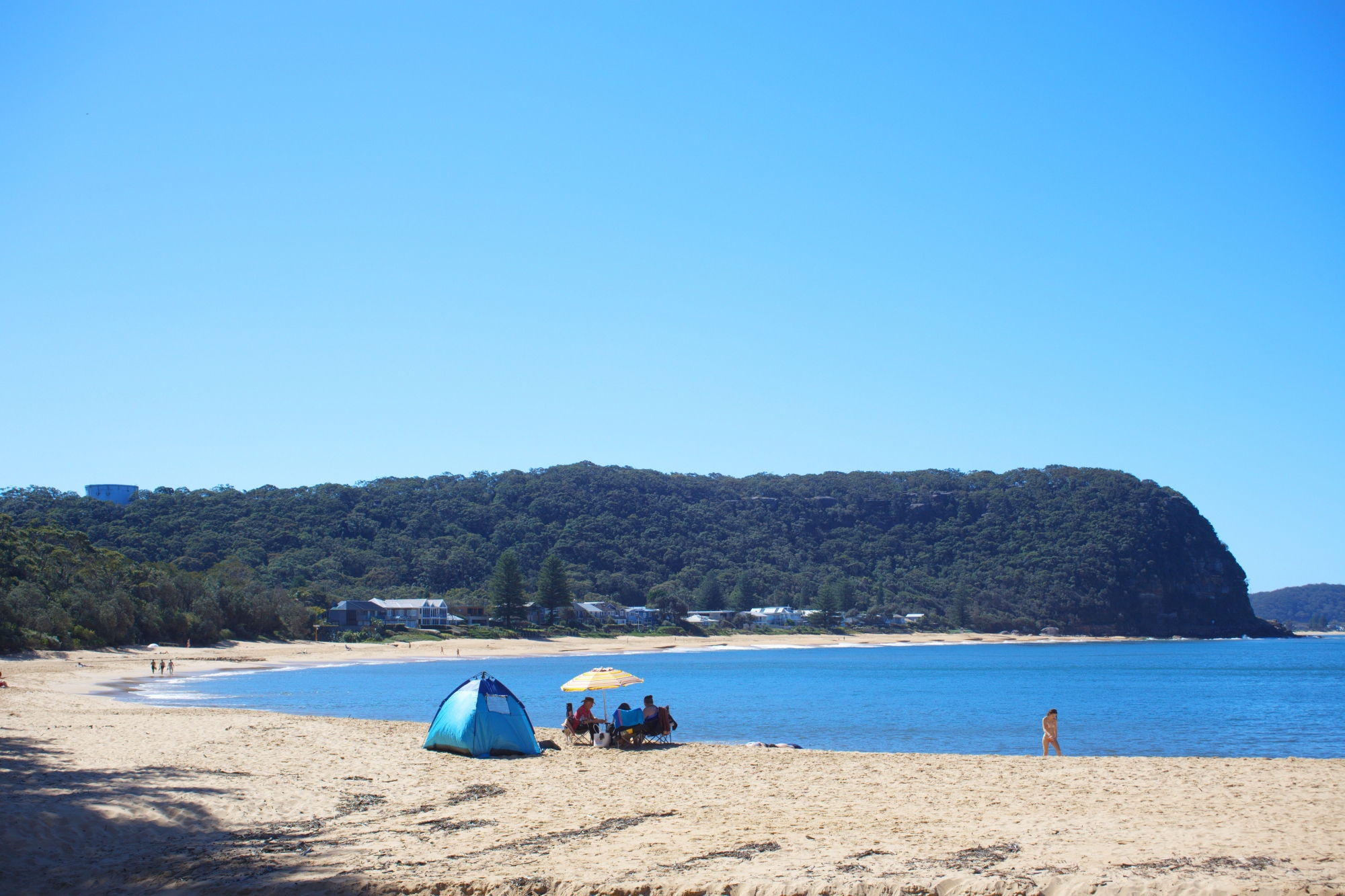 Beach tent and umbrella set up on Pearl Beach sand with the headland and clear water behind