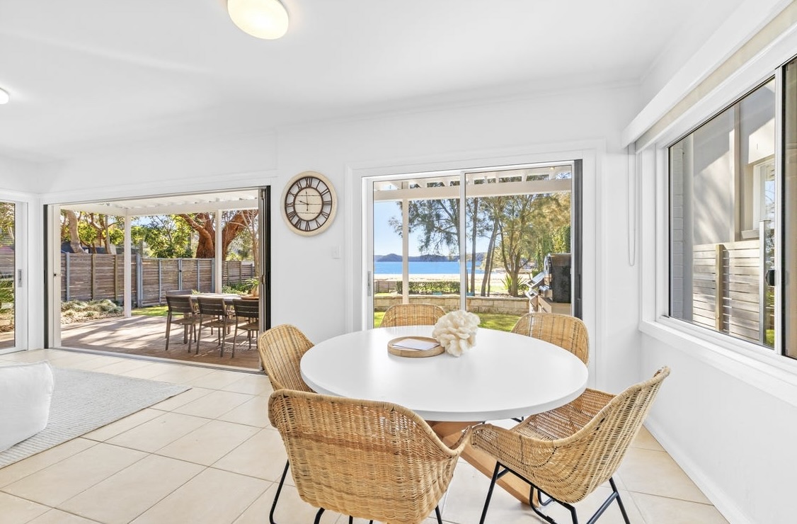 Downstairs dining nook with round white table, rattan chairs and views to the terrace and water