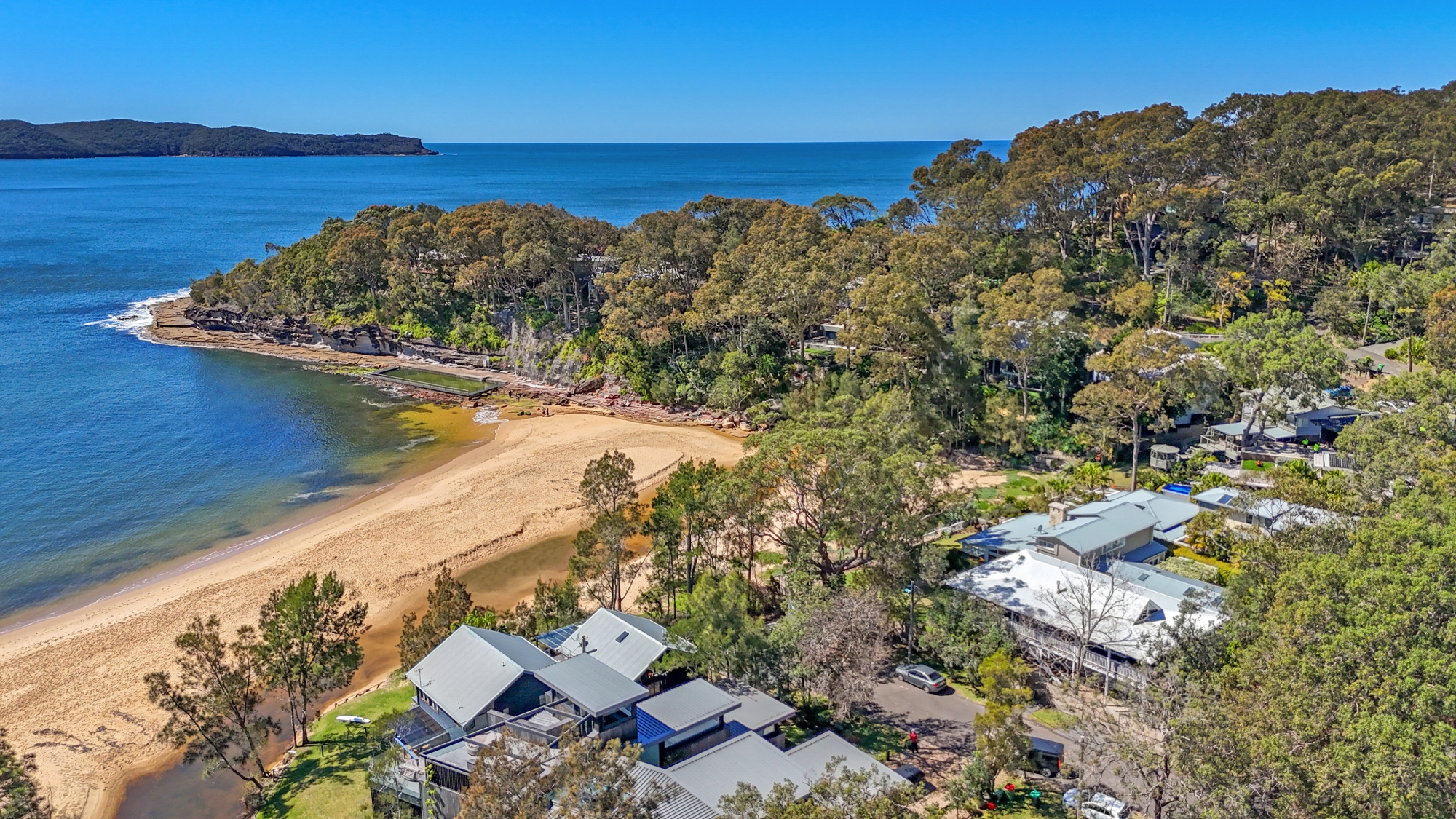 Aerial view of The Gem tucked into the bushland beside Pearl Beach, with the rock pool, sand and ocean beyond
