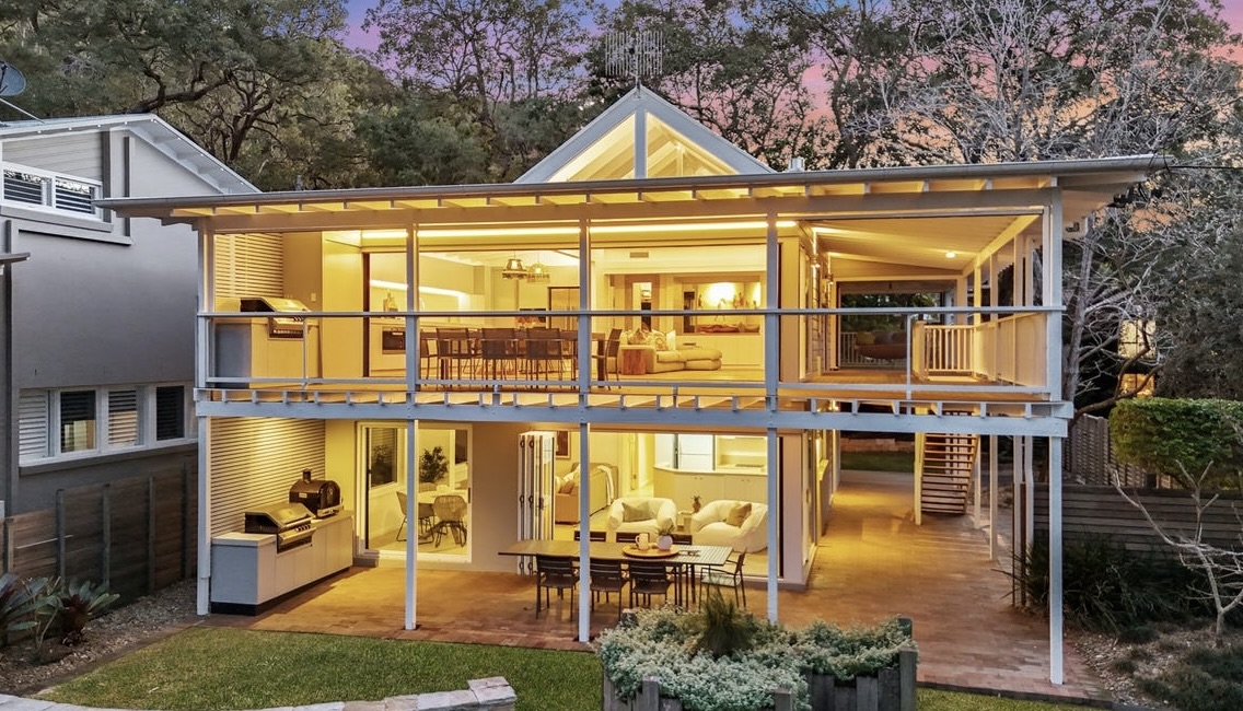 The Gem at dusk — two-storey beachfront house glowing warm against the trees, viewed from the garden