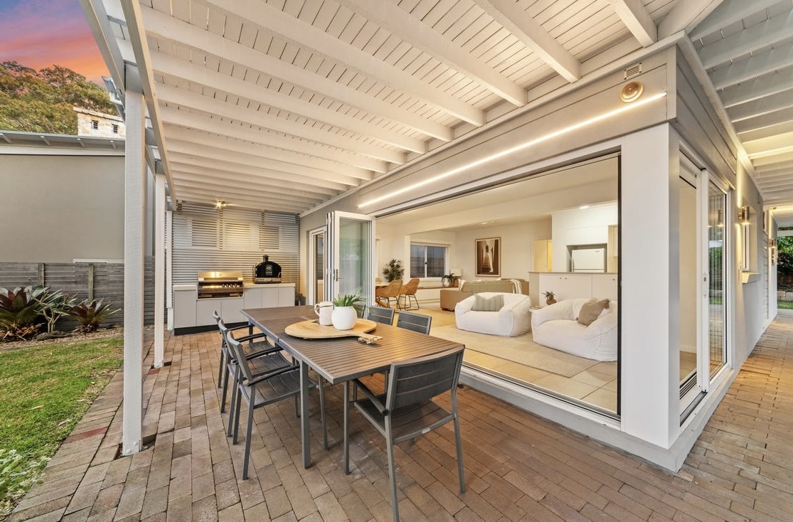 Downstairs covered terrace at dusk — outdoor dining table, BBQ area and open bifold doors leading into the living room